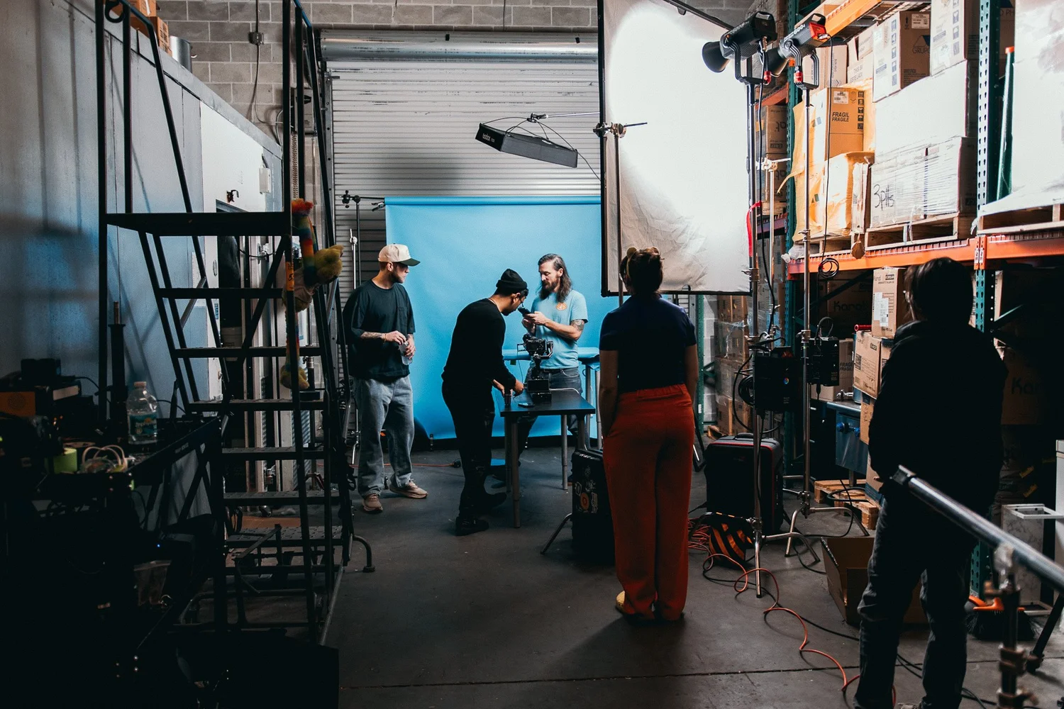 Crew around a camera on a blue paper backdrop set inside a warehouse, under an overhead light and a large diffusion frame